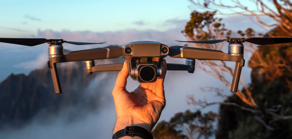 Photo of medium-sized drone with camera held in a man's hand; misty mountains in the background.