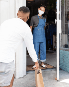 A masked young woman standing just inside the outside door of her store. A man standing about 2+ meters from the woman. with his back to the camera. He is picking up a package curb-side.
